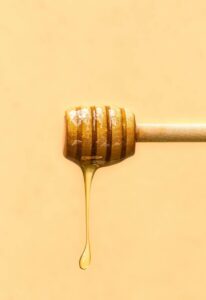 Close-up of honey drizzling from a wooden dipper against a soft yellow background.